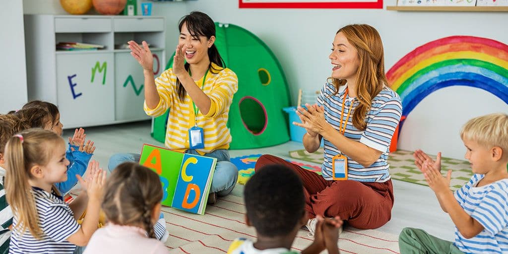 two daycare teachers sit on the floor with their students while reading them a story